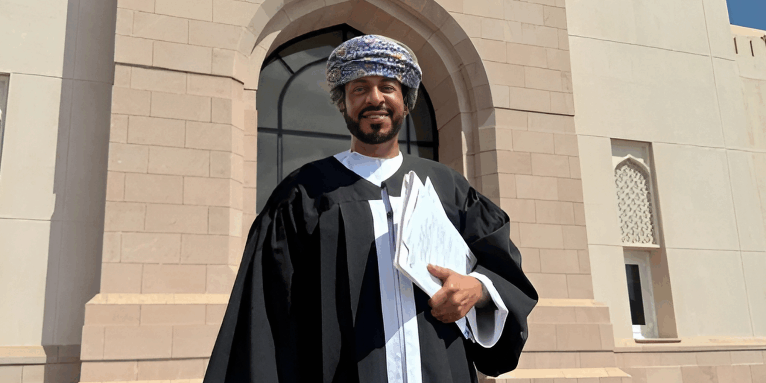 Portrait of Dr. Saleem Al Shandoudi standing in traditional legal robe in front of court-style building
